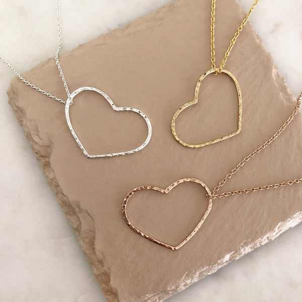 A photo of three necklaces made of dainty chains in gold, silver and rose gold featuring pendants in a large heart outline.