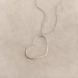 A photo of a silver necklace made of dainty chains with a large hammered heart pendant.