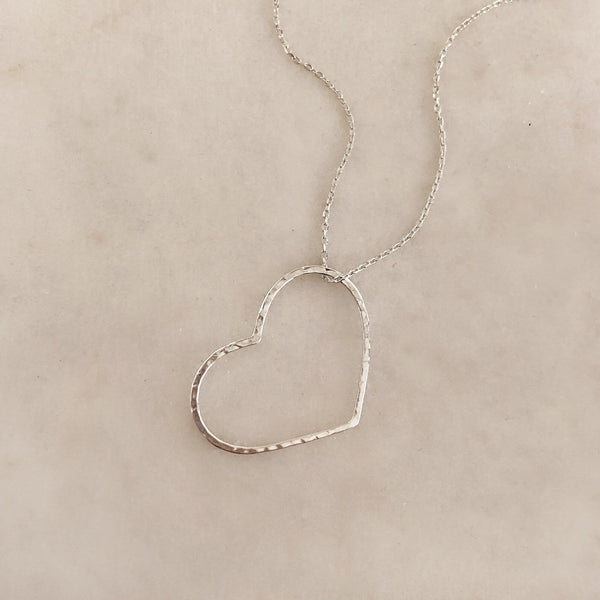 A photo of a silver necklace made of dainty chains with a large hammered heart pendant.