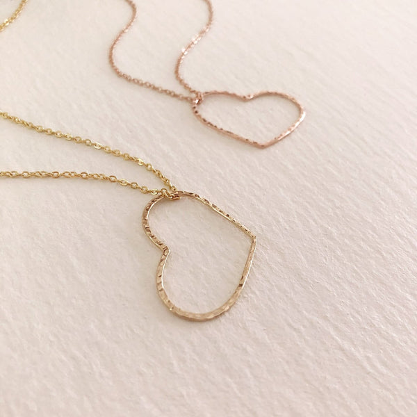 A photo of two necklaces made of dainty chains in gold and rose gold featuring pendants in a large heart outline.