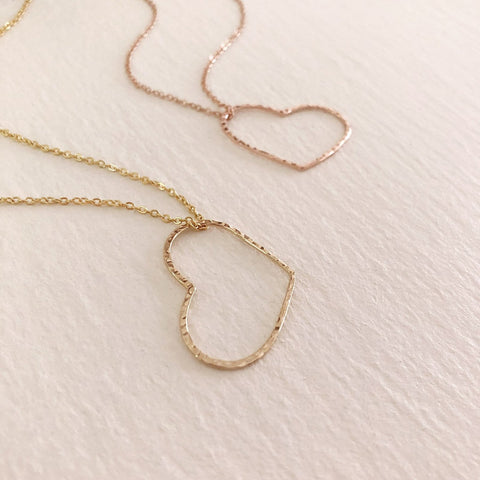 A photo of three necklaces made of dainty chains in gold, silver and rose gold featuring pendants in a large heart outline.