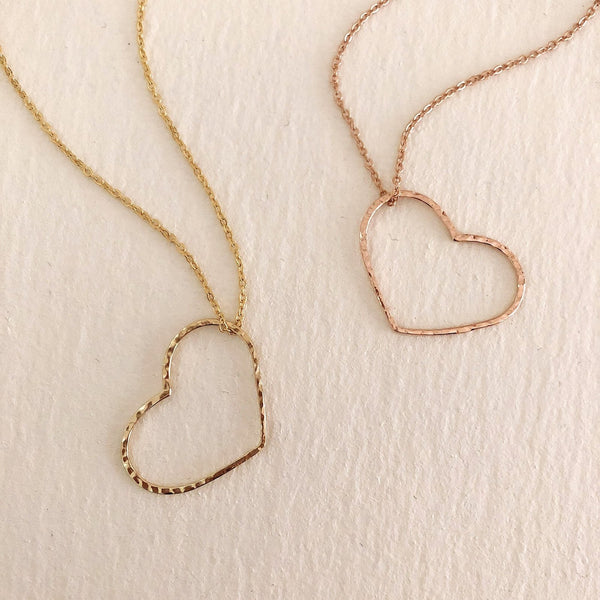 A photo of two necklaces made of dainty chains in gold and rose gold featuring pendants in a large heart outline.