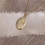 Gold necklace featuring an oval medal pendant with a design of a rose, on a dainty figaro chain.