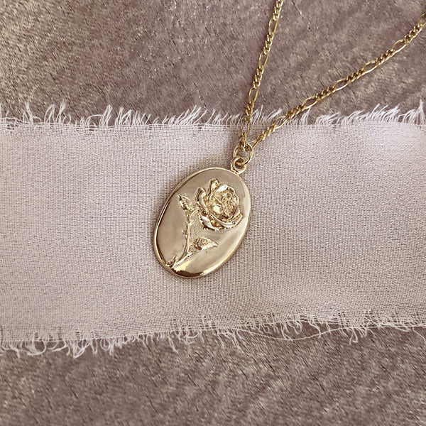 Gold necklace featuring an oval medal pendant with a design of a rose, on a dainty figaro chain.
